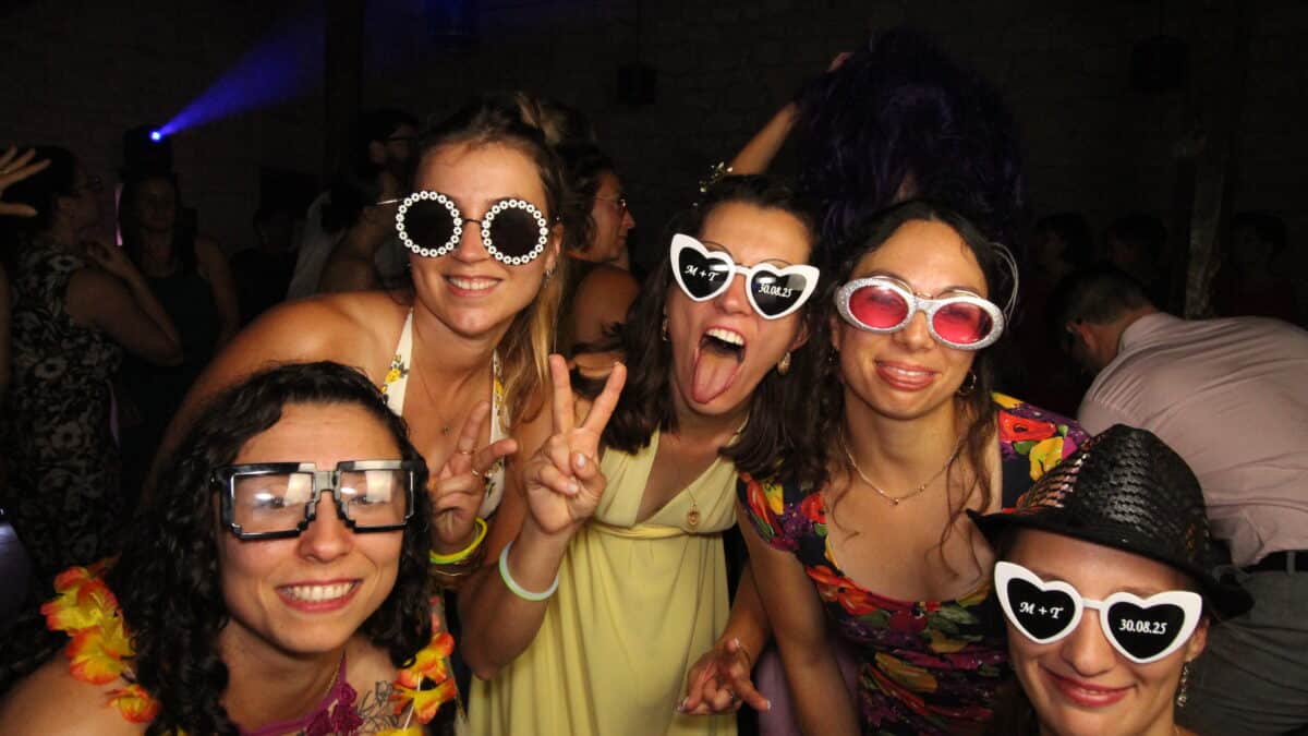 "Cinq femmes souriantes posent pour une photo lors d'une fête, portant des lunettes fantaisistes et des accessoires amusants. Elles sont habillées de robes colorées et ont l'air de s'amuser dans une atmosphère festive avec d'autres personnes à l'arrière-plan."