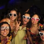 "Cinq femmes souriantes posent pour une photo lors d'une fête, portant des lunettes fantaisistes et des accessoires amusants. Elles sont habillées de robes colorées et ont l'air de s'amuser dans une atmosphère festive avec d'autres personnes à l'arrière-plan."