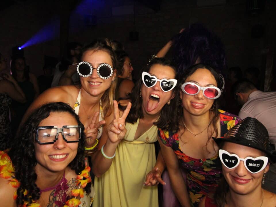 "Cinq femmes souriantes posent pour une photo lors d'une fête, portant des lunettes fantaisistes et des accessoires amusants. Elles sont habillées de robes colorées et ont l'air de s'amuser dans une atmosphère festive avec d'autres personnes à l'arrière-plan."